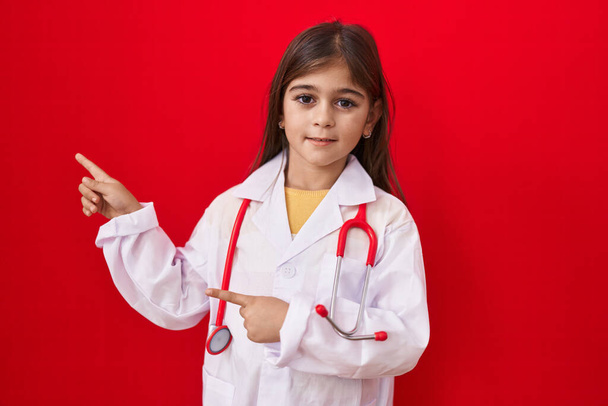 Kis spanyol lány orvos egyenruhában és sztetoszkóppal mosolyog és a kamerába néz, két kézzel és ujjal oldalra mutatva..  - Fotó, kép