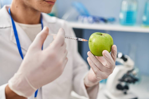 Young hispanic man wearing scientist uniform injecting on apple at laboratory - Zdjęcie, obraz