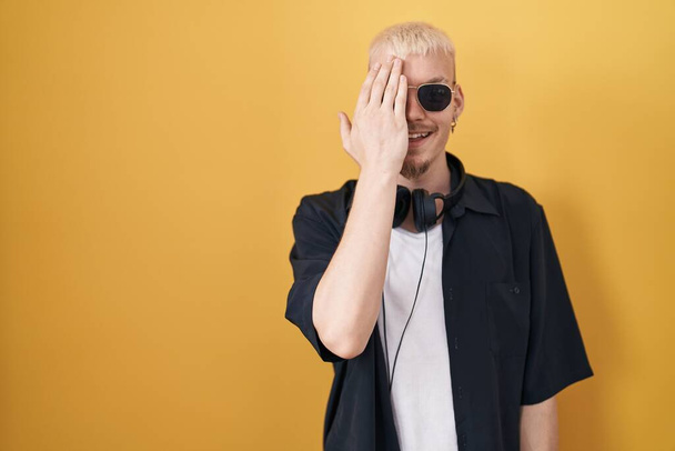 Young caucasian man wearing sunglasses standing over yellow background covering one eye with hand, confident smile on face and surprise emotion.  - Photo, Image