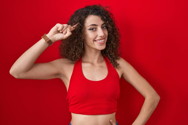 Femme hispanique avec les cheveux bouclés debout sur fond rouge souriant pointant vers la tête avec un doigt, grande idée ou pensée, bonne mémoire  - Photo, image