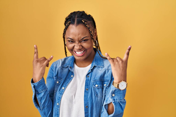 African american woman with braids standing over yellow background shouting with crazy expression doing rock symbol with hands up. music star. heavy music concept.  - Photo, image