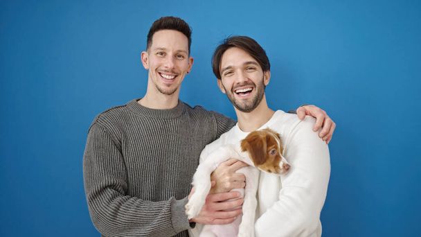 Two men couple smiling confident standing hugging dog over isolated blue background - Photo, image