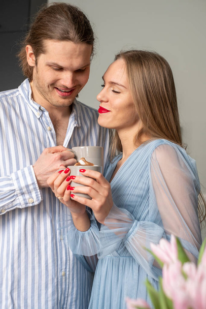 Beautiful young happy couple in light blue clothes enjoying coffee and each other's presence. Pink tulips in the corner in the foreground. Copyspace - Foto, imagen