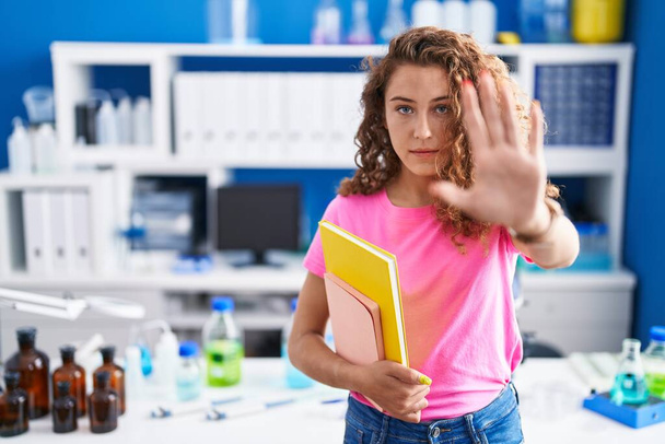 Joven estudiante caucásica en laboratorio científico con la mano abierta haciendo stop sign con expresión seria y segura, gesto de defensa  - Foto, Imagen