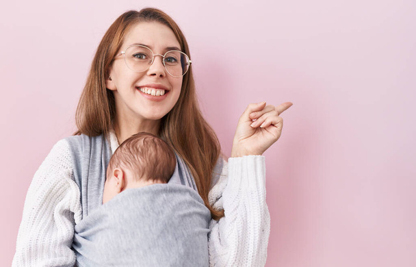 Joven mujer caucásica sosteniendo y llevando al bebé en un cabestrillo sonriendo feliz señalando con la mano y el dedo hacia un lado  - Foto, Imagen