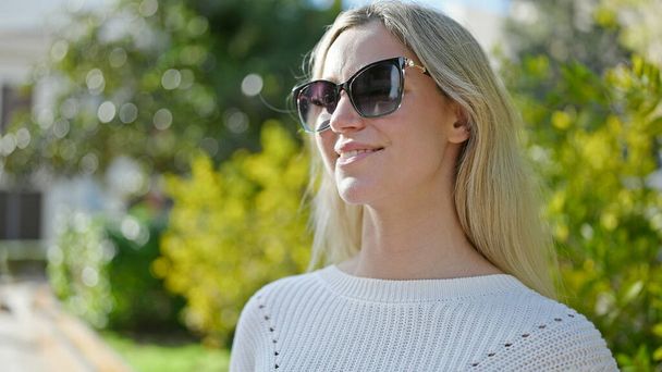 Junge blonde Frau lächelt selbstbewusst mit Sonnenbrille im Park - Foto, Bild
