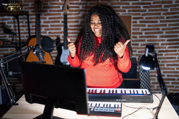 Plus rozmiar latynoska kobieta gra na fortepianie w studio muzycznym znak sukcesu robi pozytywny gest z ręki, kciuki w górę uśmiechnięty i szczęśliwy. radosna ekspresja i gest zwycięzcy.  - Zdjęcie, obraz