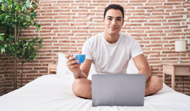 Young hispanic man using laptop drinking coffee at bedroom - Foto, imagen