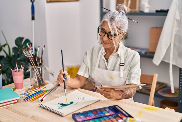 Mujer de pelo gris de mediana edad artista sonriente dibujo seguro en el cuaderno en el estudio de arte - Foto, imagen