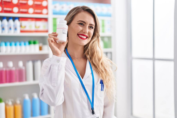 Joven hermosa mujer hispana farmacéutico sonriendo confiado sosteniendo pastillas botella en la farmacia - Foto, Imagen