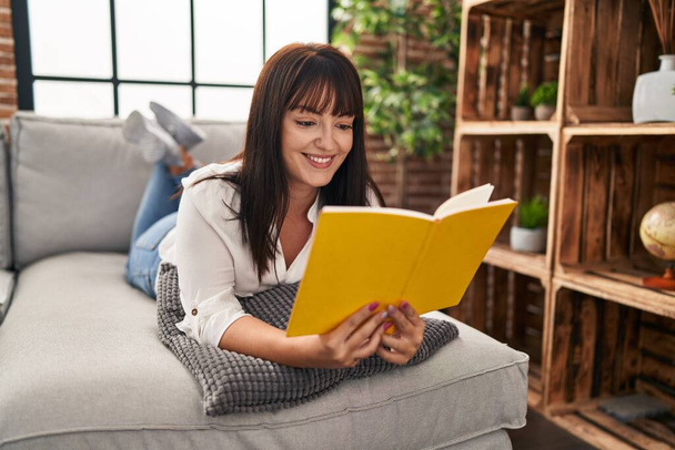 Jonge mooie Spaanse vrouw lezen boek liggend op de bank thuis - Foto, afbeelding
