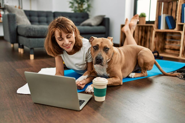 Jovem mulher caucasiana esportista sorrindo confiante usando laptop com cão em casa - Foto, Imagem