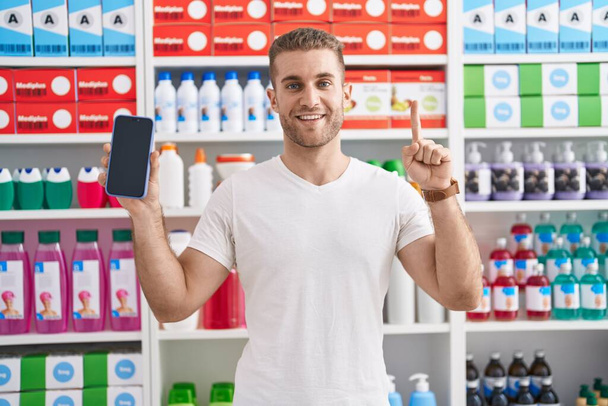 Young caucasian man working at pharmacy drugstore showing smartphone screen smiling with an idea or question pointing finger with happy face, number one  - Foto, Imagem