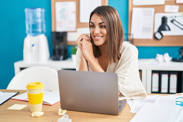 Молода красива іспанська жінка бізнесмен посміхається впевнено сидячи на столі в офісі
 - Фото, зображення