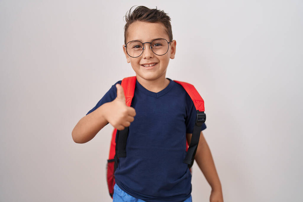 Маленький латиноамериканец в очках и студенческий рюкзак улыбается счастливым и позитивным, большой палец вверх делает отлично и знак одобрения  - Фото, изображение