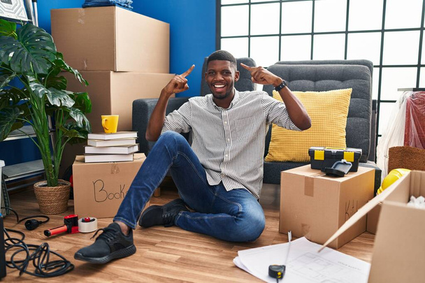 Africano americano sentado no chão na nova casa sorrindo apontando para a cabeça com as duas mãos dedo, grande ideia ou pensamento, boa memória  - Foto, Imagem