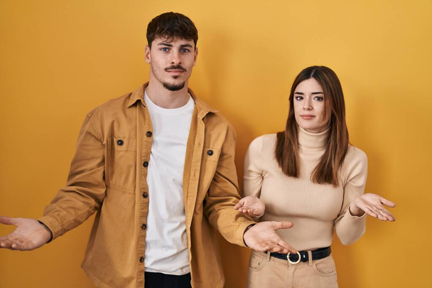 Jeune couple hispanique debout sur fond jaune désemparé et confus avec les bras ouverts, aucune idée concept.  - Photo, image