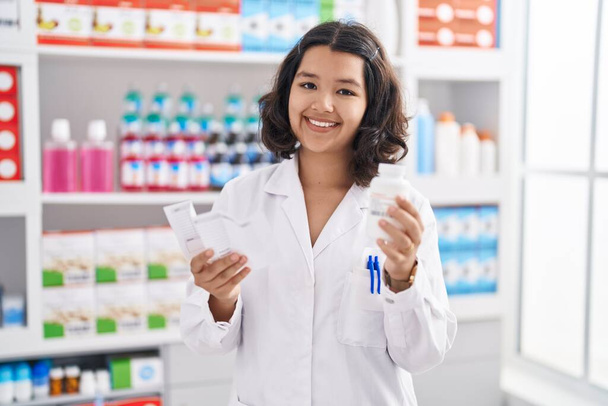 Young woman pharmacist holding pills bottle reading prescription at pharmacy - Fotoğraf, Görsel