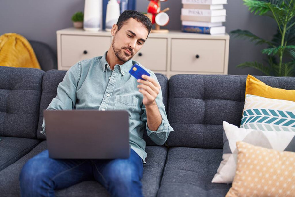 Молодий іспаномовний чоловік використовує ноутбук і кредитну картку, сидячи на дивані вдома
 - Фото, зображення