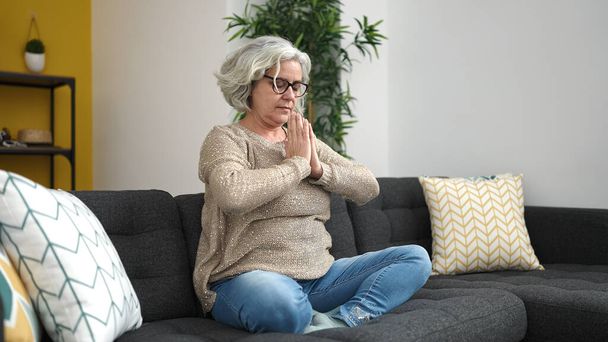 Жінка середнього віку з сірим волоссям займається вправами йоги, сидячи на дивані вдома
 - Фото, зображення