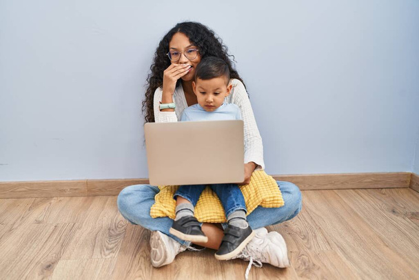 Jonge Spaanse moeder en kind met behulp van computer laptop zitten op de vloer lachen en beschaamd giechelen bedekken mond met handen, roddel en schandaal concept  - Foto, afbeelding
