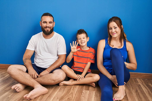Família de três sentados no chão em casa mostrando e apontando para cima com os dedos número cinco, sorrindo confiante e feliz.  - Foto, Imagem