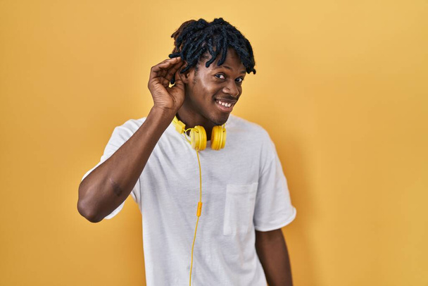 Young african man with dreadlocks standing over yellow background smiling with hand over ear listening an hearing to rumor or gossip. deafness concept.  - Foto, Bild