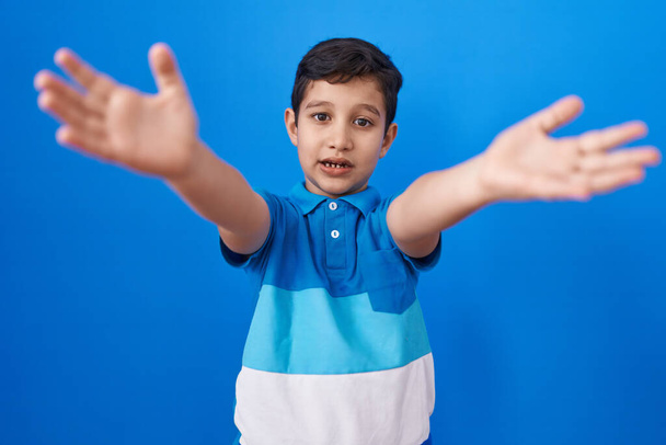 Little hispanic boy standing over blue background looking at the camera smiling with open arms for hug. cheerful expression embracing happiness.  - Zdjęcie, obraz