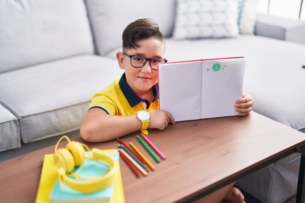 Adorable hispanic boy showing notebook with draw sitting on floor at home - Foto, afbeelding