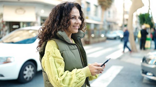 Latynoska kobieta w średnim wieku uśmiecha się pewnie używając smartfona na ulicy - Zdjęcie, obraz