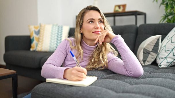 Young blonde woman writing on notebook sitting on floor thinking at home - Photo, Image