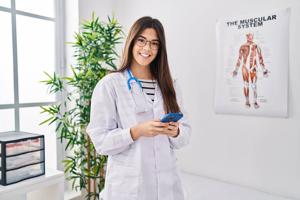 Young beautiful hispanic woman doctor smiling confident using smartphone at clinic - Photo, Image