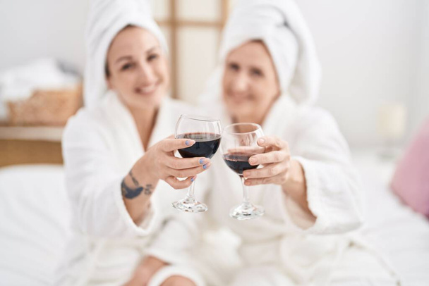 Mother and daughter wearing bathrobe drinking glass of wine at bedroom - 写真・画像