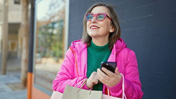 Зрелая латиноамериканка с седыми волосами улыбается, когда ходит по магазинам, используя смартфон на улице - Фото, изображение