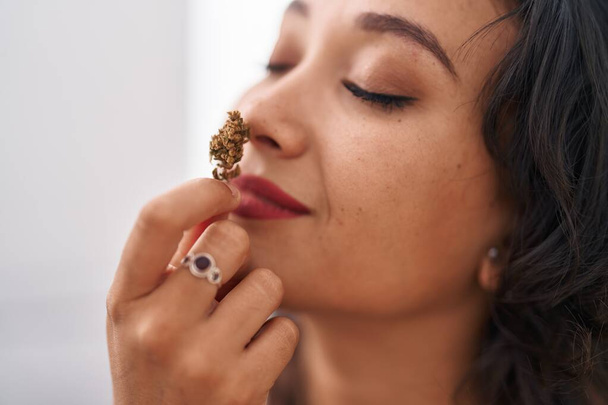 Young beautiful hispanic woman smelling marihuana plant sitting on sofa at home - Foto, Imagem