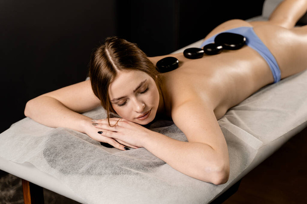 Hot stone massage therapy for relax and ease tense muscles and damaged soft tissues of body. Heated stones are placed on back of young woman - Photo, Image