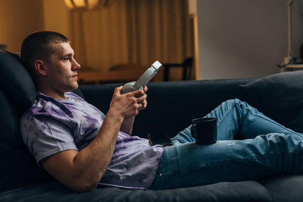 Profile view of a Caucasian man putting on headset and resting at home with a hot rink on the sofa - Foto, Bild