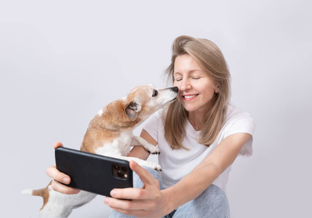 Woman and dog taking selfie. Small dog Jack Russell terrier kissing blonde woman holding using cell phone. White clothes gray solid background. Social media content creator - Фото, зображення