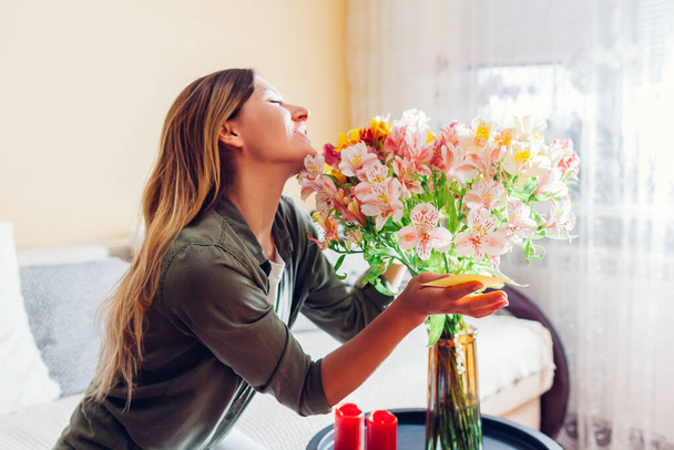 Happy young woman holds card left by boyfriend in bouquet smelling and hugging flowers at home. Surprise for holiday with note. Flower delivery - Photo, image