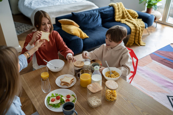 Happy smiling children brother and sister sitting at kitchen table with mom enjoying healthy breakfast together, mother and two kids siblings eating in morning at talking, family bonding over mealtime - Photo, Image