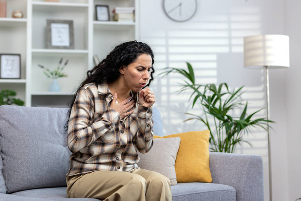 Kobieta przeziębiona i chora kaszel siedzi na kanapie w domu, Latynoska kobieta w salonie z przeziębieniem i grypą. - Zdjęcie, obraz