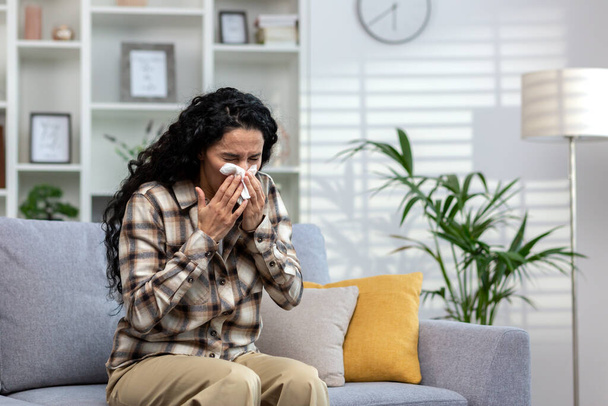 Woman with allergy sneezes sitting on sofa in living room, Latin American sick woman wipes nose with tissue at home. - Fotoğraf, Görsel
