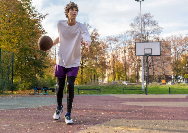 Cute young teenager in t shirt with a ball plays basketball on court. Teenager running in the stadium. Sports, hobby, active lifestyle for boys - 写真・画像
