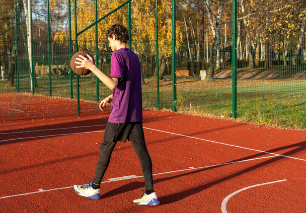 Netter junger Teenager im T-Shirt mit Ball spielt Basketball auf dem Platz. Teenager beim Laufen im Stadion. Sport, Hobby, aktiver Lebensstil für Jungen - Foto, Bild