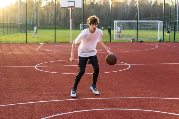 Милый молодой подросток в футболке с мячом играет в баскетбол на корте. Подросток бегает по стадиону. Спорт, хобби, активный образ жизни для мальчиков - Фото, изображение