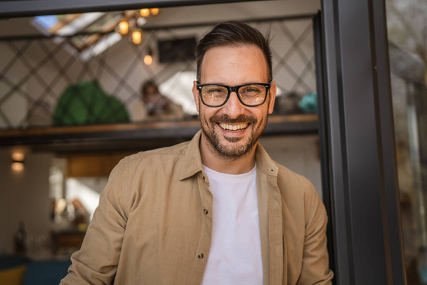 One man adult caucasian male with beard and eyeglasses stand on balcony at door in day happy smile hold cup of coffee copy space morning daily routine concept - Fotografie, Obrázek