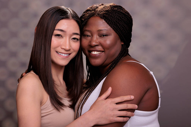 Dos mujeres diversas abrazándose demostrando confianza, amor y autocuidado. Hermosa africana americana y asiática jóvenes novias sonriendo, abrazando juntos y mirando a la cámara - Foto, Imagen