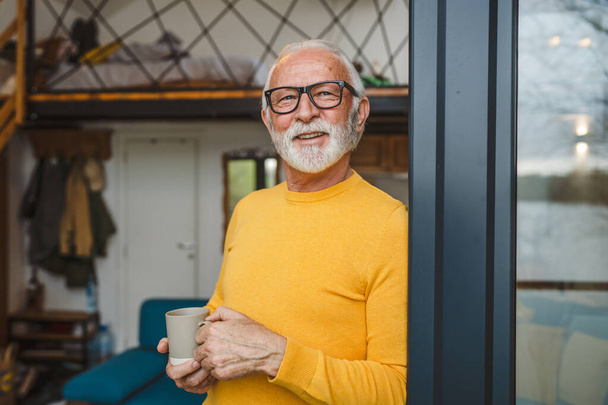 髭と眼鏡を持つ一人の男性高齢者の白人男性が一日のドアのバルコニーに立つ幸せな笑顔は、コーヒーコピースペースのカップを保持します毎日ルーチンのコンセプト - 写真・画像