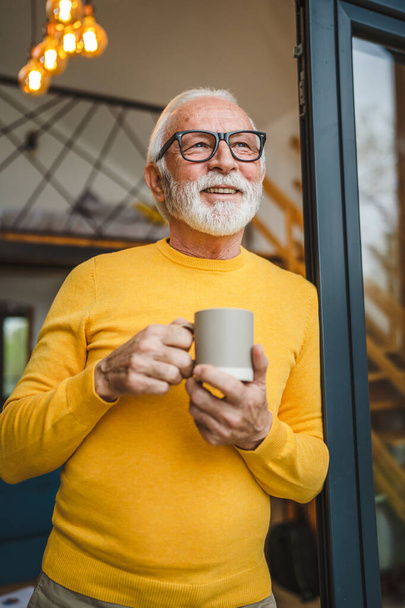 Egy férfi vezető kaukázusi férfi szakállal és szemüveggel áll az erkélyen az ajtóban nappal boldog mosoly tart csésze kávé másolás tér reggel napi rutin koncepció - Fotó, kép