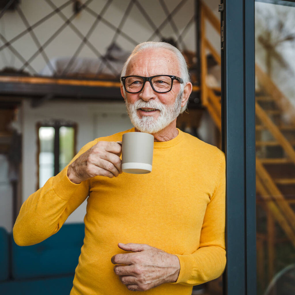 Un uomo anziano maschio caucasico con barba e occhiali stare sul balcone alla porta in giorno sorriso felice tenere la tazza di caffè copia spazio mattina concetto di routine quotidiana - Foto, immagini